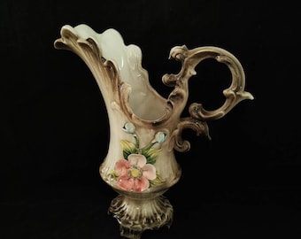 May include: A vintage ceramic pitcher with a scalloped rim and ornate handle. The pitcher is primarily cream-colored with brown accents and a floral design featuring a pink flower and blue buds. The base has a decorative, textured design.