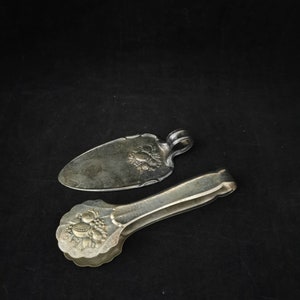 Peut inclure: Un ensemble de deux ustensiles de service argentés. L'un est un serveur à gâteau avec une poignée décorative et l'autre est une pince à épiler avec un motif floral.