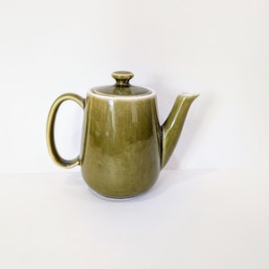 Puede incluir: Tetera de cerámica verde oliva con asa curva y boquilla. La tetera tiene una tapa con un pequeño pomo en la parte superior. La tetera es de color liso y tiene un acabado brillante. Es un artículo de menaje.