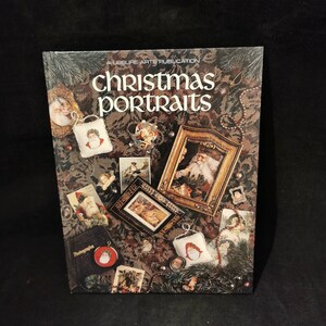Puede incluir: Un libro titulado "Christmas Portraits" con una cubierta decorativa que presenta varias imágenes navideñas, incluidos retratos enmarcados de Papá Noel y otros adornos festivos. La cubierta tiene un fondo oscuro y estampado.