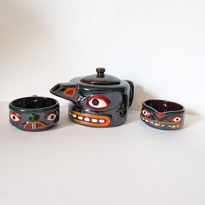 Może przedstawiać: Ceramiczny czajnik i dwie filiżanki z ozdobnym wzorem twarzy. Czajnik jest brązowy z zielonymi, czerwonymi, pomarańczowymi i białymi detalami. Filiżanki są brązowe z zielonymi, czerwonymi i białymi detalami.