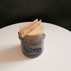 Könnte beinhalten: Eine graue Keramik-Gurkenglas mit einem Holzdeckel und einem blau-weißen Design. Das Glas trägt den Schriftzug "Gurken Apfel".