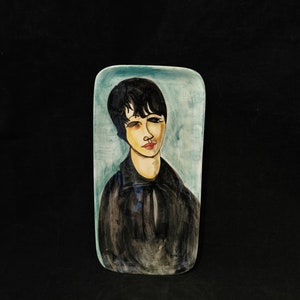 May include: A rectangular ceramic plate featuring a painted portrait of a person with short black hair. The person is wearing a black shirt and has a light blue background.