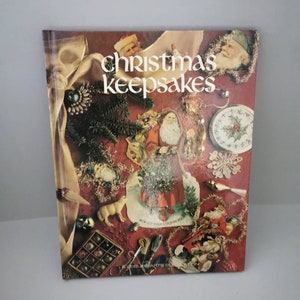 Puede incluir: Un libro vintage titulado "Christmas Keepsakes" con una portada roja y un collage de adornos navideños, decoraciones y recortes de papel. El libro presenta una variedad de elementos festivos, incluyendo una figura de Papá Noel, un reno y una media.