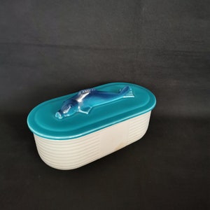 Pode incluir: Uma travessa oval de cerâmica com tampa turquesa e escultura de peixe azul. A base branca apresenta detalhes canelados horizontais. Ideal para servir ou armazenar alimentos.