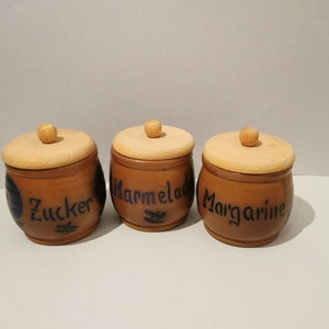 May include: Three wooden jars with lids, each labelled with a different food item: "Zucker", "Marmelad", and "Margarine".