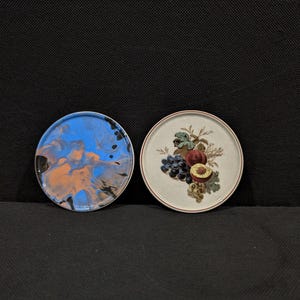 Puede incluir: Dos posavasos redondos. Uno presenta un diseño abstracto azul, naranja y negro. El otro posavasos tiene un fondo crema con una ilustración de frutas de uvas, melocotones y hojas. Ambos posavasos tienen un borde oscuro.