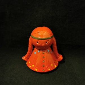 Könnte beinhalten: Eine Keramikfigur einer jungen Frau in einem roten Kleid, einem grünen Stirnband und gelben und blauen Akzenten. Die Figur hat ein verspieltes und fantasievolles Design.