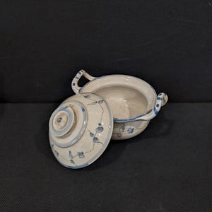 Könnte beinhalten: Eine weiße Keramik-Servierschale mit Deckel, verziert mit blauen Blumenmustern. Die Schale hat zwei Griffe und einen passenden Deckel, beide mit zarten blauen Designs. Der Deckel ist teilweise geöffnet und zeigt das Innere der Schale.