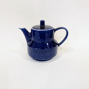 Puede incluir: Una tetera de cerámica azul oscuro con un patrón geométrico en la parte inferior. Tiene un asa curva, un pico y una tapa con un pequeño pomo. Un borde blanco acentúa la tapa. Diseño clásico.