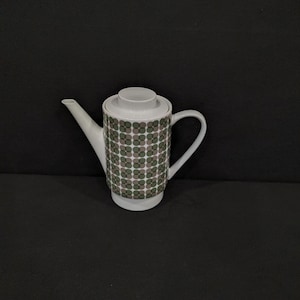 Op de afbeelding: Witte keramische theepot met een deksel en handvat. De body van de theepot heeft een herhalend patroon van groene en grijze cirkels. De tuit is lang en slank. De theepot staat tegen een donkere achtergrond.