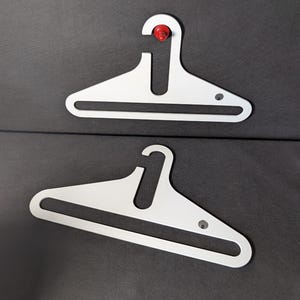 Puede incluir: Dos perchas blancas con un diseño minimalista. Cada percha tiene un botón rojo en la parte superior y un pequeño detalle circular en la parte inferior. Las perchas están sobre un fondo gris oscuro.