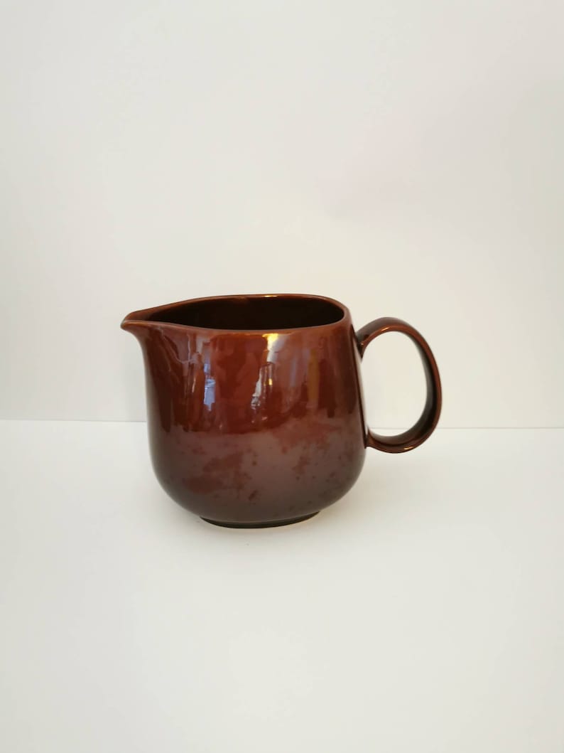 May include: A brown ceramic pitcher with a rounded body and a handle. The pitcher has a glossy finish.