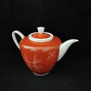 Può includere: Una teiera in ceramica con corpo arrotondato e manico, beccuccio e coperchio bianchi. La teiera è principalmente arancione con linee e punti decorativi bianchi. Il coperchio ha un piccolo pomello bianco.