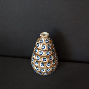 Può includere: Un piccolo vaso in ceramica a forma di goccia con un collo stretto. Il vaso è marrone con un motivo di cerchi blu e verdi. I cerchi sono disposti in un motivo ripetuto sulla superficie del vaso.