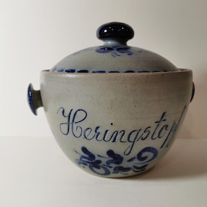 Op de afbeelding: Een grijze steengoed pot met een blauw deksel en blauwe bloemmotieven. De pot heeft een blauwe inscriptie met de tekst "Heringstopf".