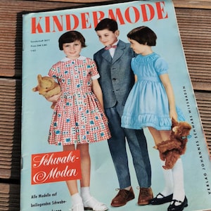 Pode incluir: Capa de revista vintage "Kindermode" com três crianças. A capa tem um fundo azul claro com texto vermelho. As crianças usam roupas vintage, incluindo vestidos e um terno. A revista é de 1962.