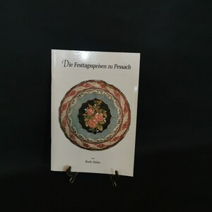 May include: A book titled "Die Festtagsspeisen zu Pessach" with a decorative plate illustration on the cover. The plate features a floral design in the center, surrounded by Hebrew text and a red and blue border. The book is displayed on a black stand against a black background.