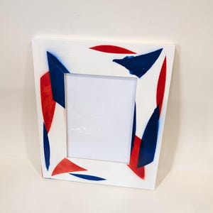 May include: White picture frame with abstract red and blue paint strokes. The frame has a rectangular opening for a photo. The design is modern and artistic, suitable for displaying artwork or photographs.