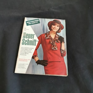 Könnte beinhalten: Ein Vintage-Magazin-Cover mit einer Frau in rotem Anzug und Hut. Der Magazintitel lautet "Neuer Schnitt" mit deutschem Text. Das Cover enthält auch den Text "Neue Herbstmode für jeden Typ" und "10 Oktober 1964".