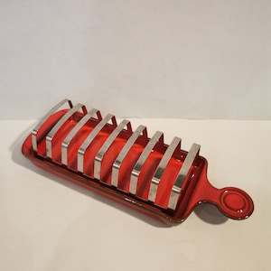 Puede incluir: Portacuchillos para pan de cerámica roja con una rejilla metálica para sujetar las rebanadas de pan. El portacuchillos tiene un asa en el lateral.