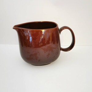 May include: A brown ceramic pitcher with a rounded body and a handle. The pitcher is glazed and has a smooth, glossy finish.