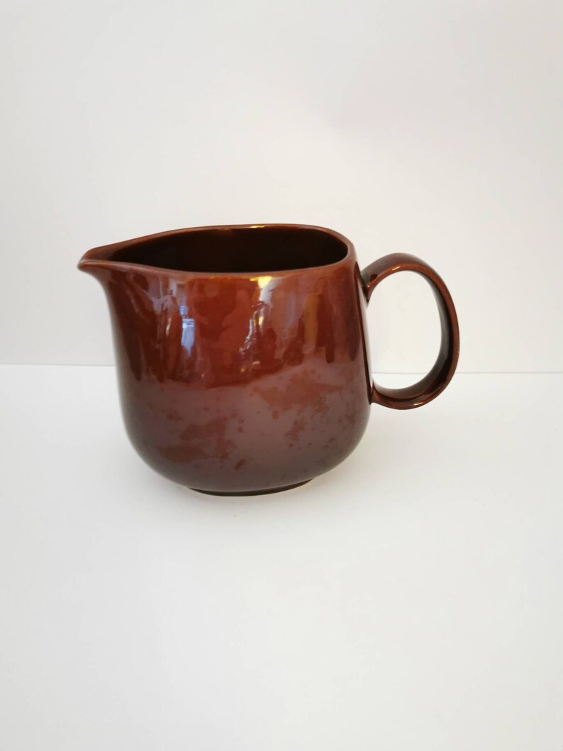 May include: A brown ceramic pitcher with a rounded body and a handle. The pitcher is glazed and has a smooth, glossy finish.