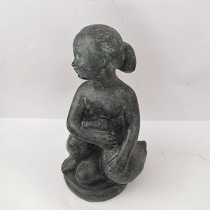 Puede incluir: Una escultura de bronce verde de una niña sentada en una roca. La niña tiene los brazos alrededor de las rodillas y la cabeza inclinada hacia abajo.