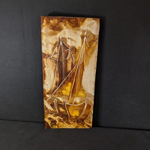Könnte beinhalten: Ein rechteckiges Holzkunstwerk mit zwei Segelbooten. Die Segelboote sind in Sepiatönen gehalten, mit detaillierten Linien und Schattierungen. Das Kunstwerk hat ein rustikales, handwerkliches Aussehen mit sichtbarer Holzmaserung.