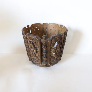 Peut inclure: Un pot en métal décoratif de couleur bronze avec un design orné et texturé. Le pot a une forme de panier avec un bord supérieur festonné et des motifs en relief détaillés, notamment des motifs floraux et géométriques. Le pot est probablement vintage ou antique.