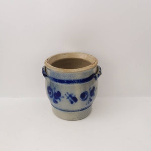 Puede incluir: Una pequeña maceta de cerámica blanca con diseños florales azules y dos asas. La maceta está esmaltada y tiene un aspecto rústico.