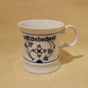 Puede incluir: Taza de cerámica blanca con asa y diseño floral azul decorativo. La taza tiene la palabra "Klönbecher" en escritura azul. La taza tiene una base acampanada y un asa curva.