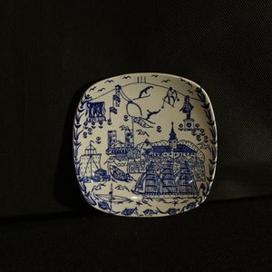 Puede incluir: Un plato cuadrado de cerámica blanca con un diseño azul y blanco que representa una ciudad con barcos y la palabra "OSLO" en el centro.