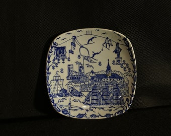 May include: A white ceramic square plate with a blue and white design depicting a cityscape with ships and the word "OSLO" in the center.
