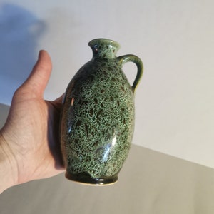 Puede incluir: Un pequeño jarrón de cerámica verde con un esmalte moteado y un asa marrón.