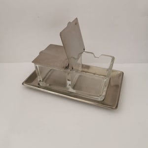 Peut inclure: Un distributeur de sucre en argent et en verre avec un couvercle à charnière. Le distributeur a deux compartiments et repose sur un plateau en argent.