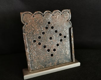 May include: A decorative metal object with a square, perforated design and a scalloped top edge. The object has a rustic, aged appearance with a silver base and is displayed against a dark background.
