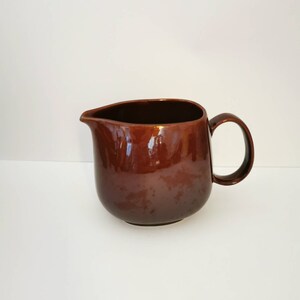 May include: A brown ceramic pitcher with a rounded body and a handle. The pitcher has a glossy finish.