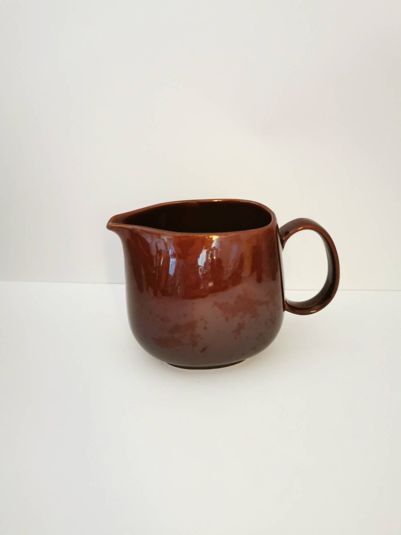 May include: A brown ceramic pitcher with a rounded body and a handle. The pitcher has a glossy finish.