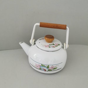 Puede incluir: Tetera de esmalte blanco con un diseño floral en rosa y azul. La tetera tiene un asa y un pomo de tapa de madera, y un asa de metal blanco. Un artículo de cocina clásico.