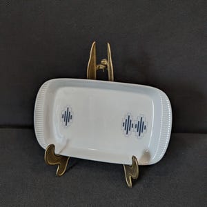 May include: A white rectangular ceramic dish with a blue geometric pattern, displayed on a gold-coloured metal stand. The dish has a ribbed edge and is approximately 20 cm long. The geometric pattern is repeated twice on the dish.