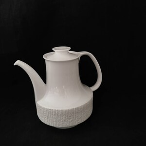 May include: White ceramic teapot with a textured base and a lid. The teapot has a spout and a handle.