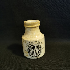 Puede incluir: Un tarro de mostaza de cerámica blanca con una etiqueta negra que dice "Ce Pot de Moutarde doit porter la capsule. Marque Deposee." y un círculo negro con el texto "Grey-Poupon" en su interior.