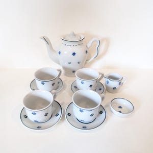 Puede incluir: Juego de té de porcelana blanca con detalles florales azules. El conjunto incluye una tetera, cuatro tazas con platillos, una jarra para crema y un cuenco pequeño. Los bordes son azules.
