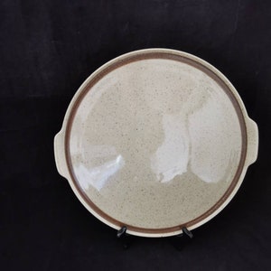 Könnte beinhalten: Eine große, runde, beige Servierplatte aus Keramik mit braunem Rand und zwei Griffen. Die Platte hat eine gesprenkelte Oberfläche.