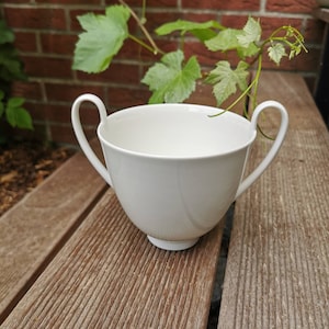 Könnte beinhalten: Eine weiße Porzellantasse mit zwei Henkeln. Die Tasse steht auf einer Holzoberfläche.