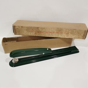 Puede incluir: Una grapadora de metal verde con una palanca plateada y una base plateada. La grapadora está en su caja original, que es marrón y tiene el texto "HEFTAPPARAT NOVUS-LANG 24/6" impreso en ella.