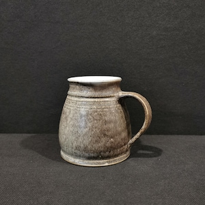 Könnte beinhalten: Eine handgefertigte Keramiktasse mit rundem Körper und stabilem Henkel. Die Tasse hat eine gesprenkelte, erdig-braune Glasur mit weißem Innenraum. Die Tasse ist etwa 10 cm hoch, perfekt für Kaffee oder Tee.