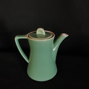 Puede incluir: Una tetera de cerámica de color verde azulado con tapa y asa. La tetera está sobre un fondo negro.