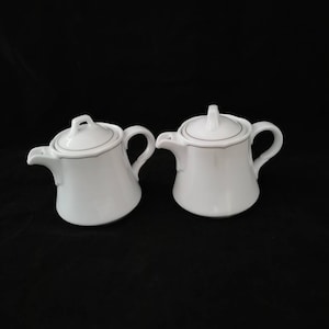 Könnte beinhalten: Zwei weiße Teekannen aus Keramik mit schwarzem Besatz und Deckeln. Die Teekannen stehen auf einem schwarzen Hintergrund.